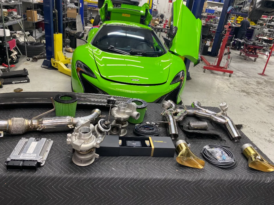 Green McLaren supercar in a shop with an array of performance upgrade parts including turbos, exhaust, and ECU laid out.
