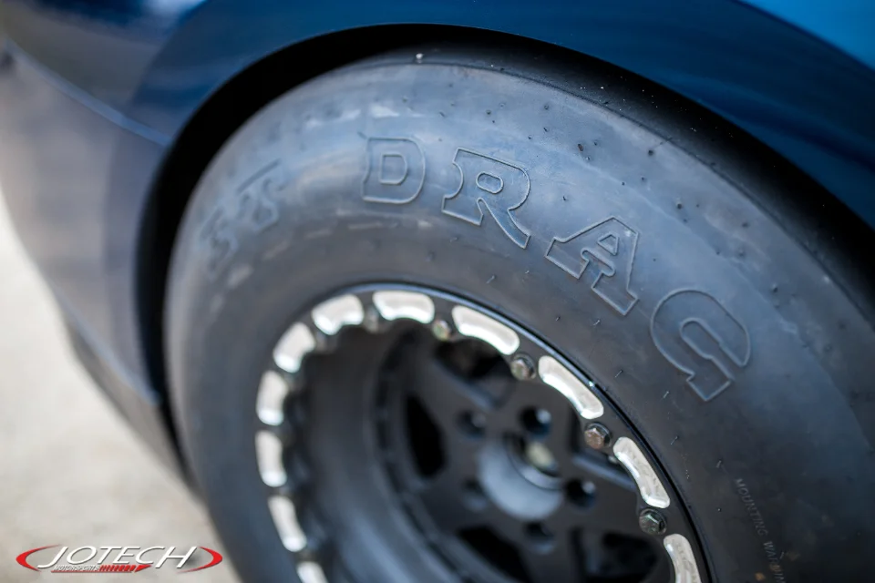 Close-up of a drag tire on a black beadlock wheel mounted on a blue performance car, with Jotech branding.