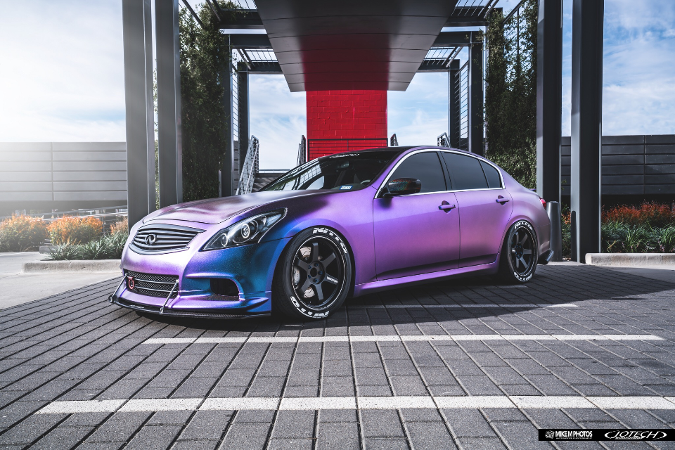A custom purple and blue iridescent wrapped Infiniti G37 sedan with aftermarket wheels and a front splitter parked in a modern setting.