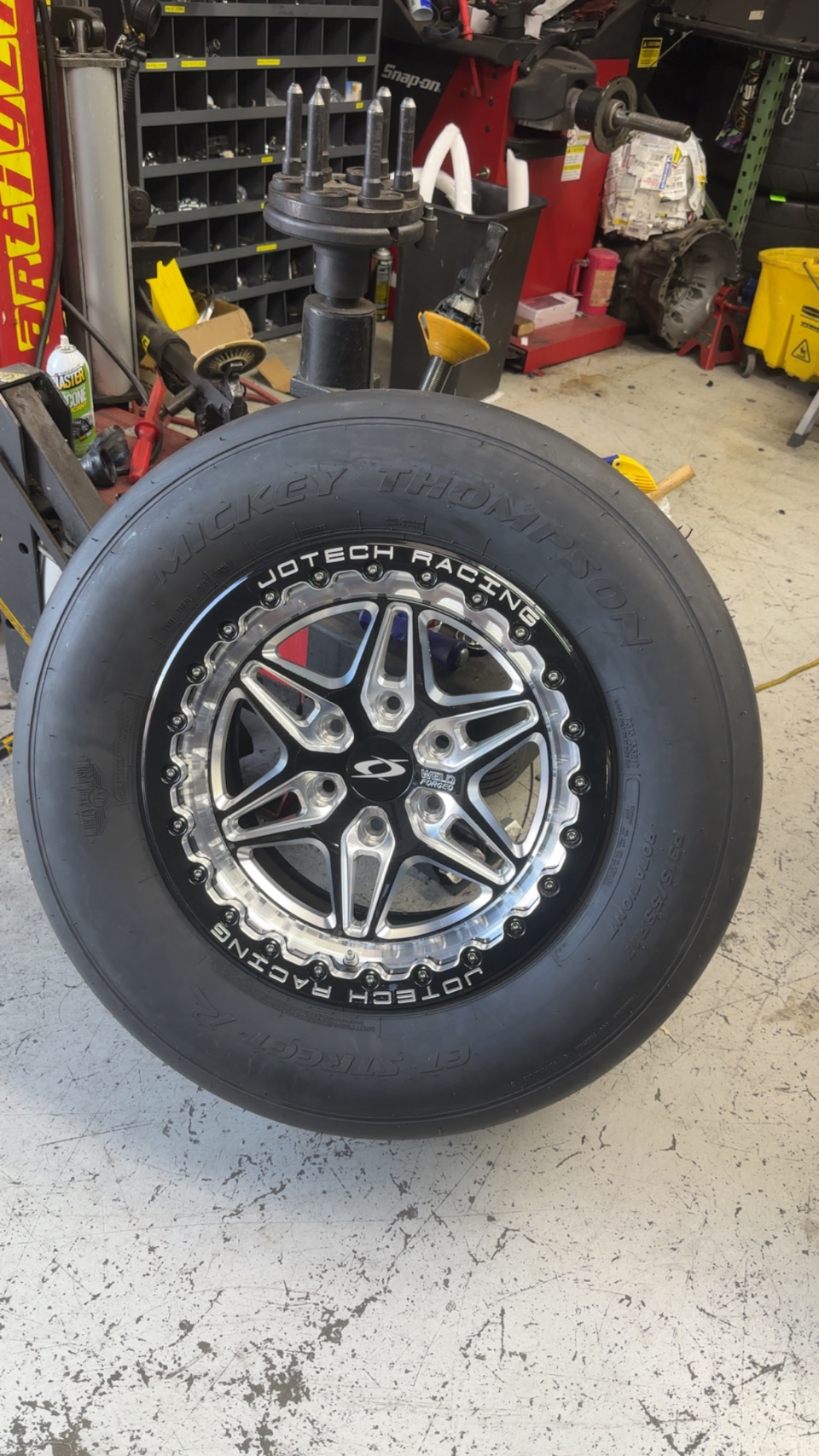 A Mickey Thompson drag radial tire mounted on a black and polished Weld Forged wheel with Jotech Racing branding, in a shop.
