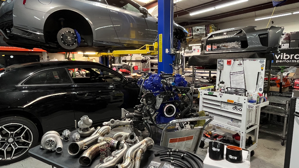 A grey Nissan GT-R on a lift and a black coupe in a performance tuning shop, with a blue engine block and various turbo and exhaust parts on a table.