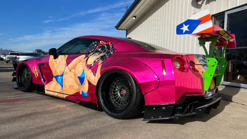 A vibrant pink Nissan GT-R with a widebody kit, anime-style wrap, large green rear wing, and aftermarket wheels.