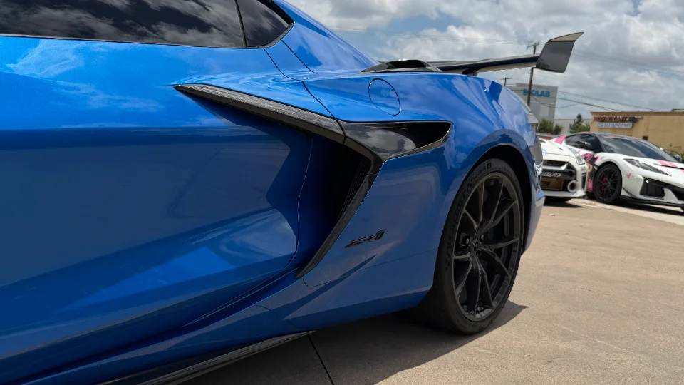 A blue Chevrolet Corvette C8 Z06 with black wheels and a rear wing, parked outdoors with other cars in the background.