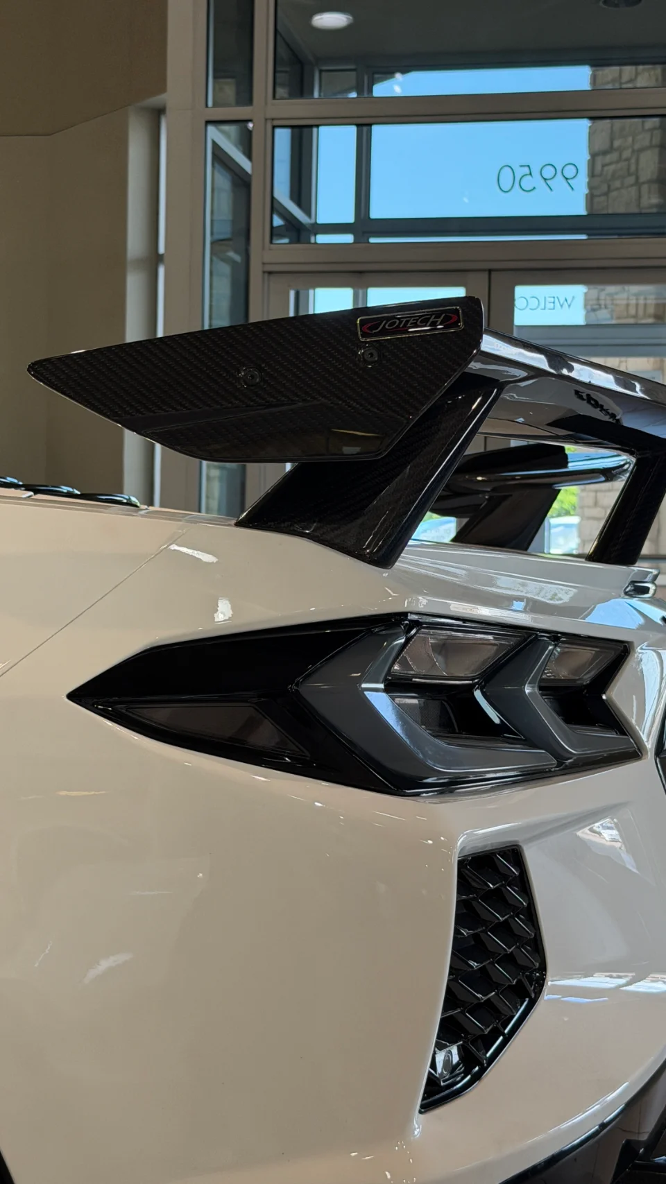 Close-up of a white Corvette's rear with a Jotech carbon fiber wing and LED taillights in a showroom.