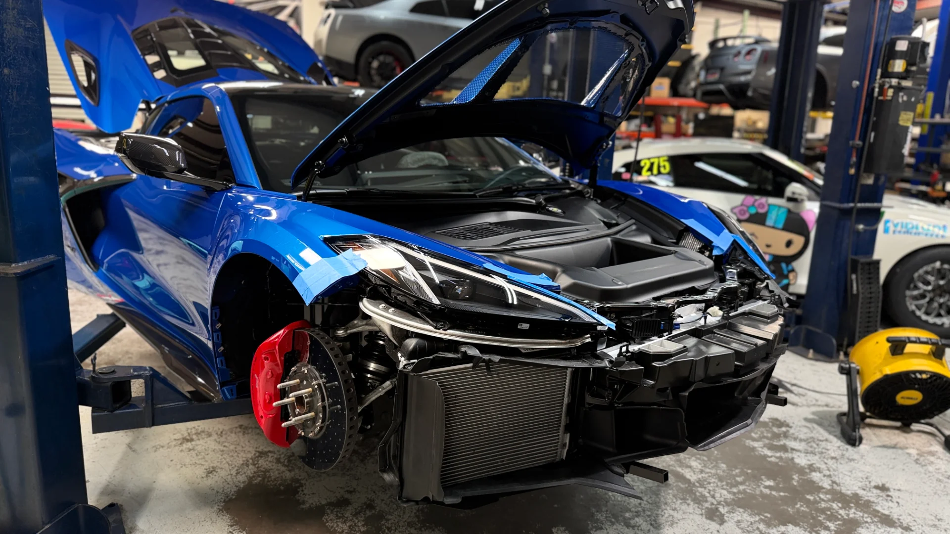 A blue Chevrolet C8 Corvette on a lift in a performance shop with its front bumper removed and hood open, revealing the engine and front end components.