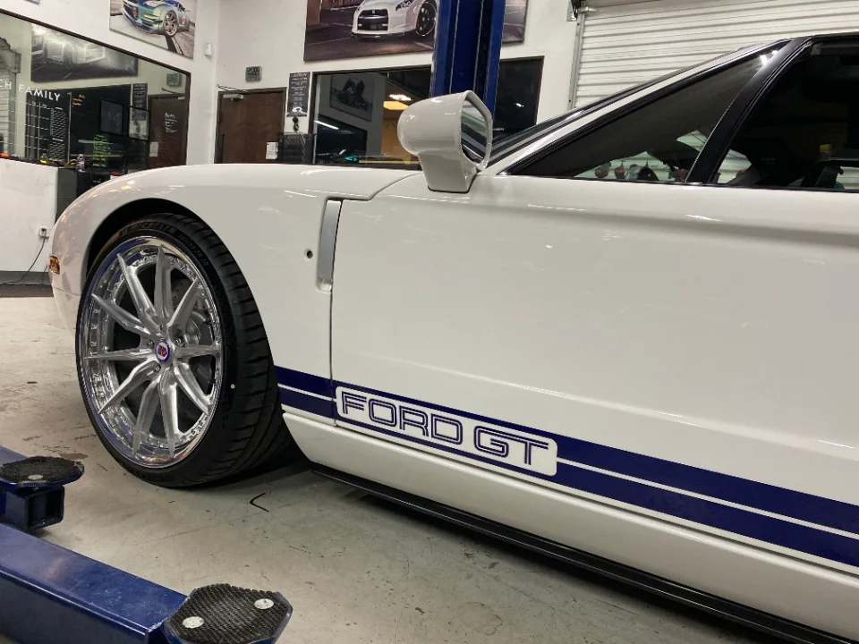 A white Ford GT with blue racing stripes and aftermarket chrome wheels on a lift in a performance shop.