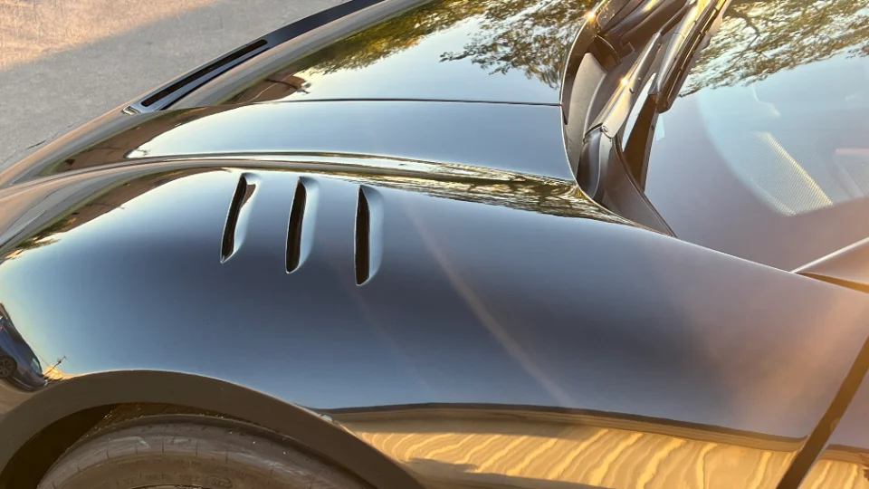 Close-up of a black car's shiny fender with three vertical vents, reflecting trees and sunlight.
