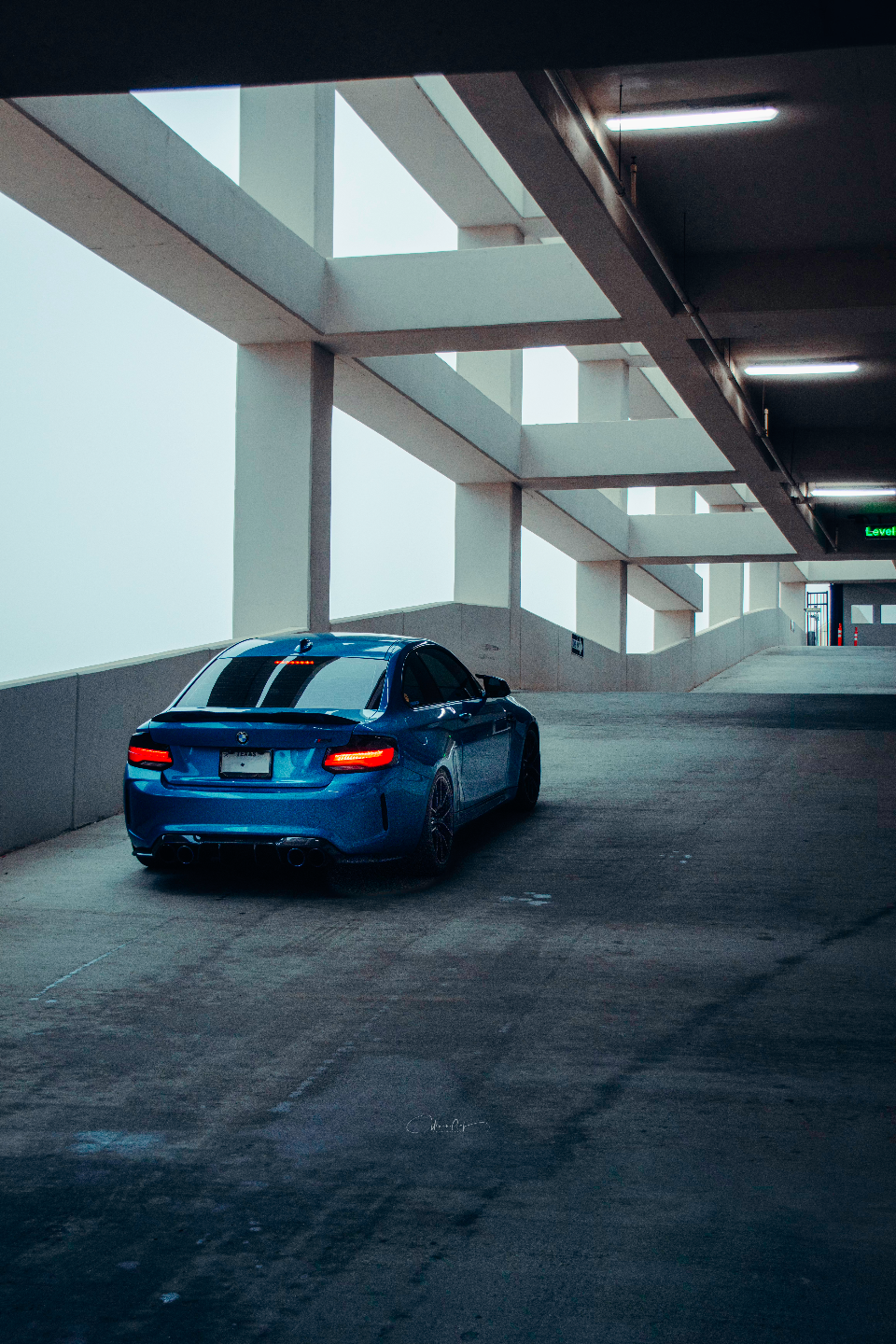A blue BMW M2 sports car parked in a concrete parking garage, viewed from the rear with its taillights illuminated.