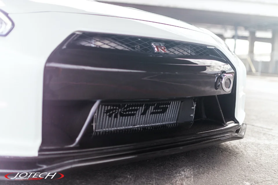 Close-up of a white Nissan GT-R R35 front bumper featuring an aftermarket intercooler, carbon fiber lip, and tow hook.