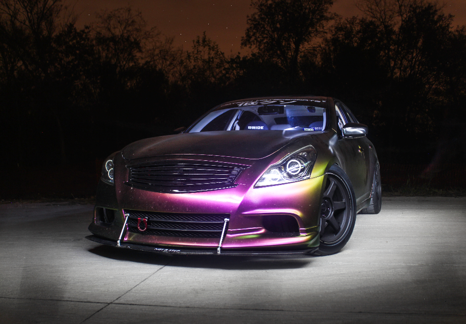 A custom Infiniti G37 coupe with a chameleon wrap, aftermarket wheels, and a front splitter, professionally lit at night.