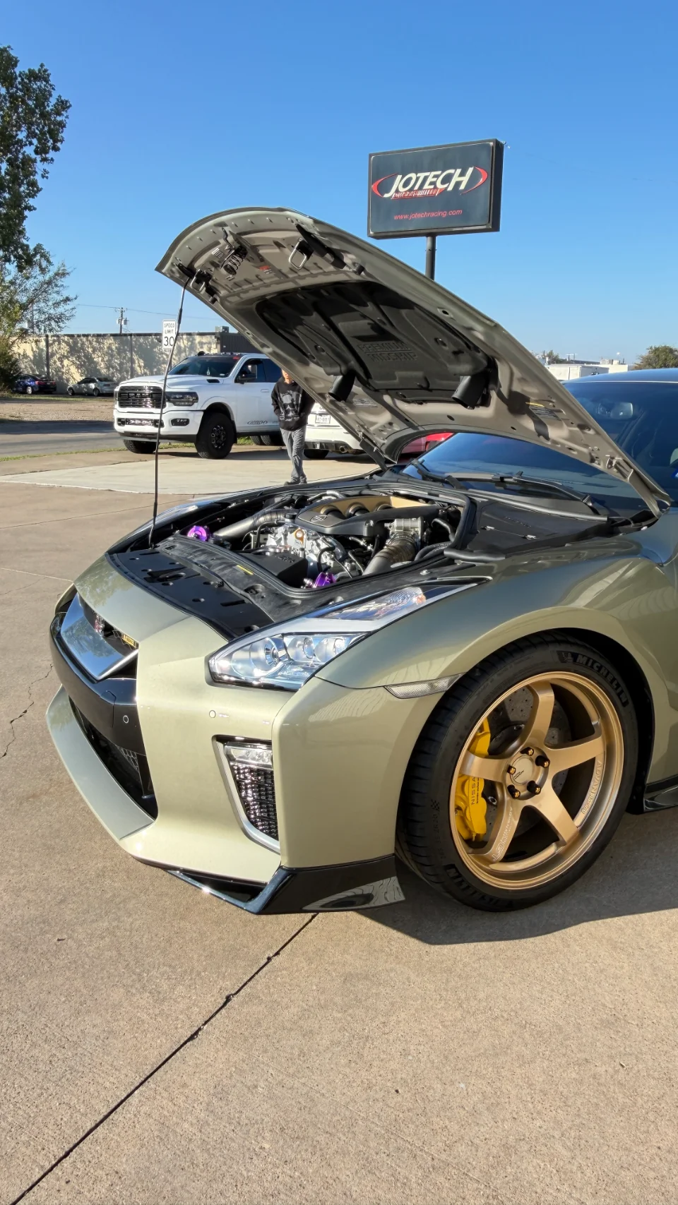 A gold Nissan GT-R with its hood open, revealing a modified engine bay, parked outside Jotech Motorsports with gold wheels and yellow brake calipers.