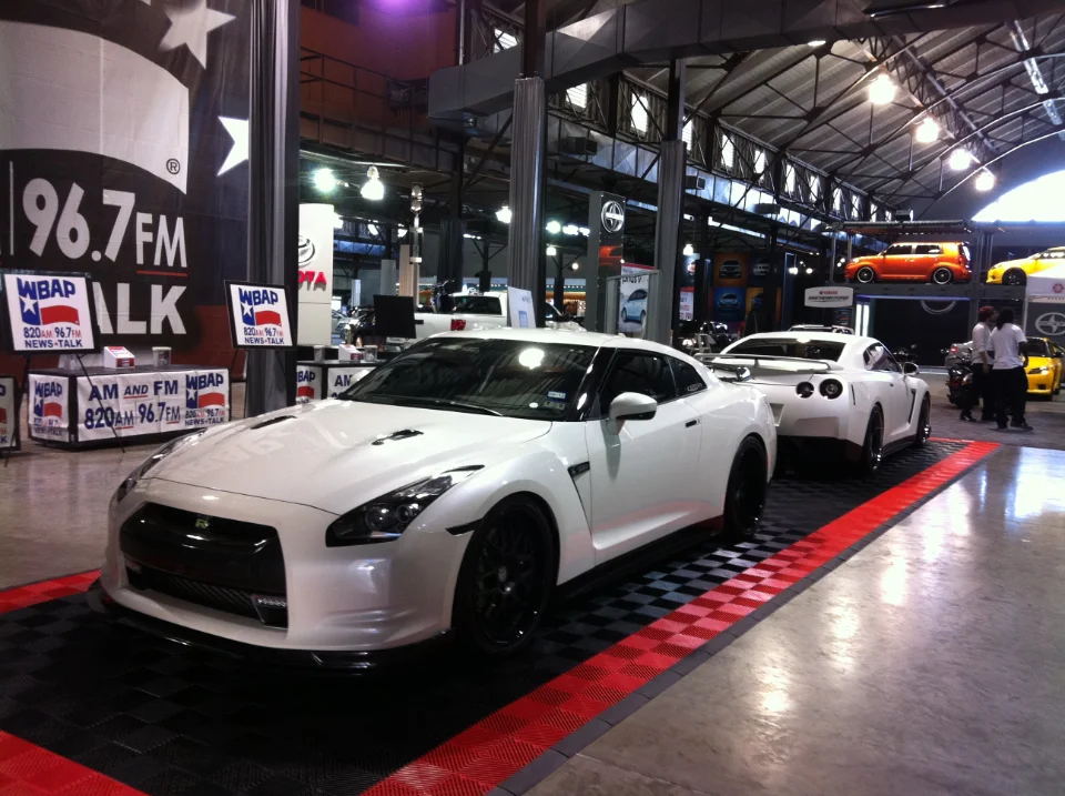 Two white Nissan GT-Rs on display at an indoor car show or event, with radio station banners in the background.