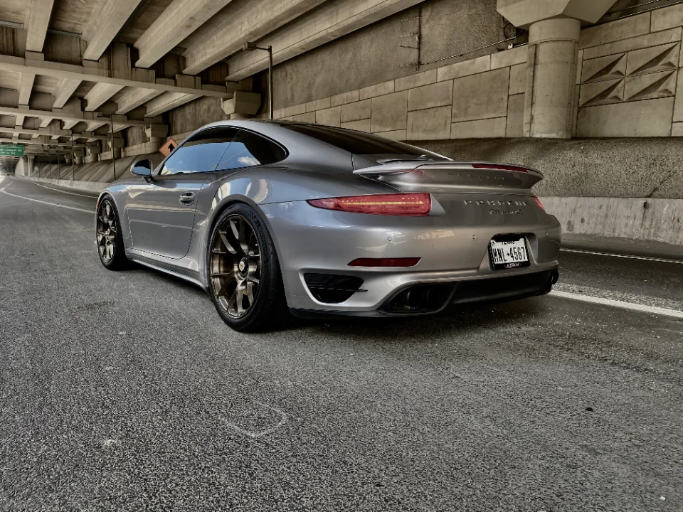 A silver Porsche 911 Turbo S with aftermarket wheels parked under a concrete overpass.