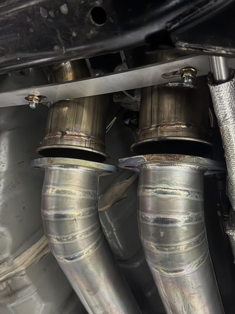 Close-up view of custom stainless steel exhaust downpipes installed under a performance car, showing welds and flanges.