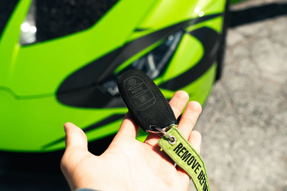 A hand holding a McLaren car key with a "Remove Before Flight" keychain in front of a bright green McLaren supercar.