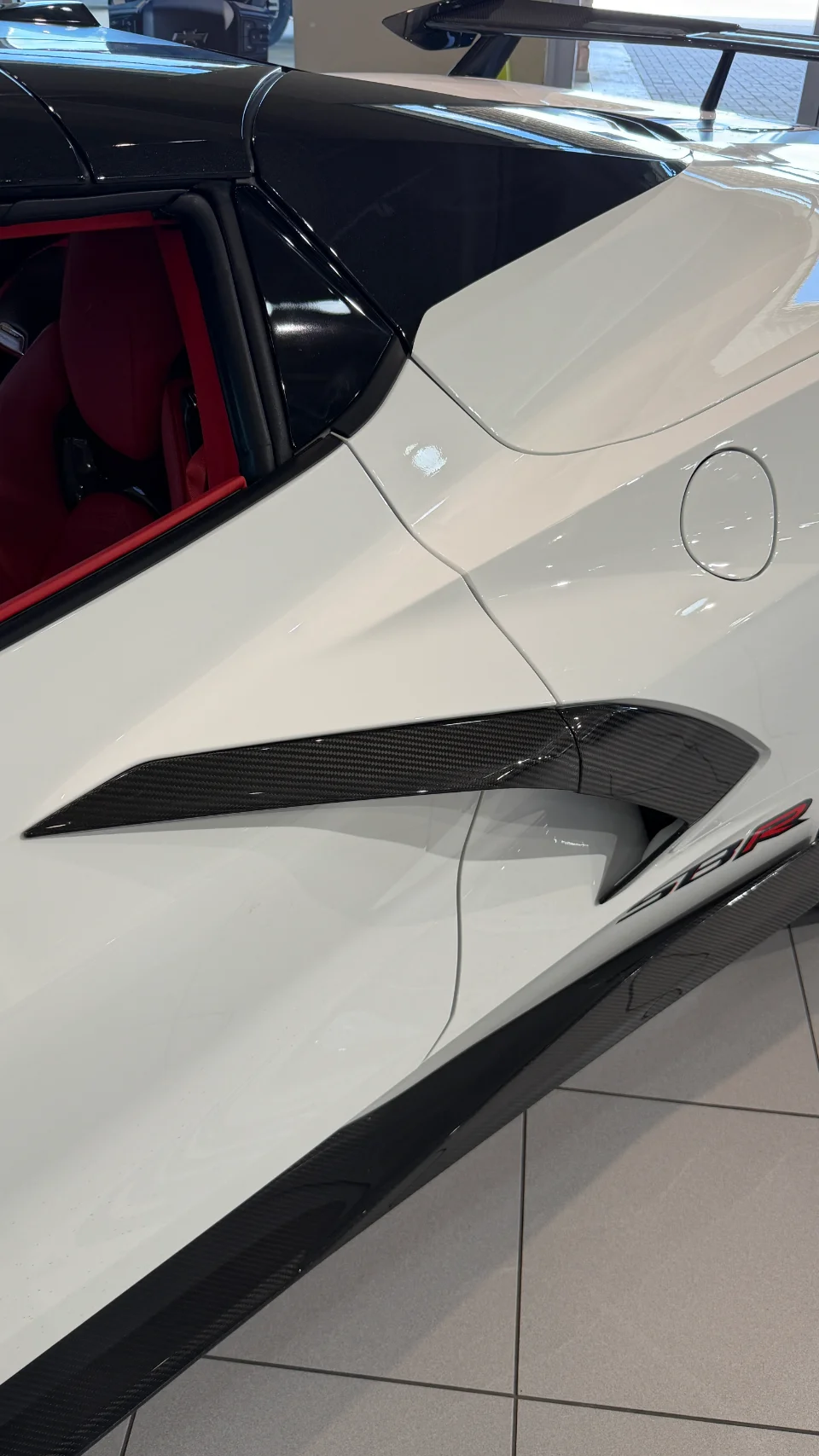 A white Corvette C8 with carbon fiber side intake and rocker panel, showcasing exterior aerodynamic details and red interior.