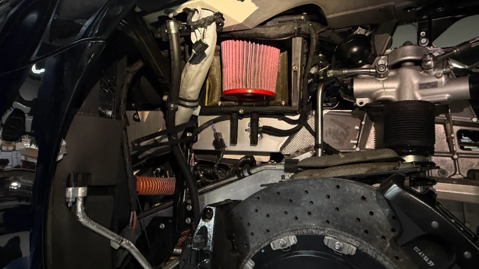 Close-up of a McLaren's engine bay, showcasing an aftermarket red air filter, performance brake rotor, and suspension components.