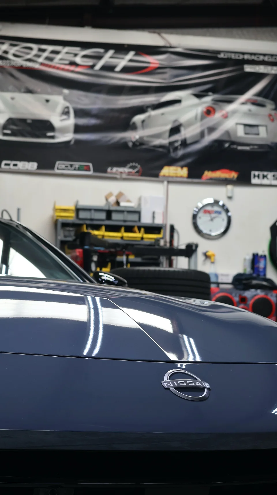 Front view of a blue Nissan Z parked inside the Jotech Motorsports shop with tuning brand banners in the background.