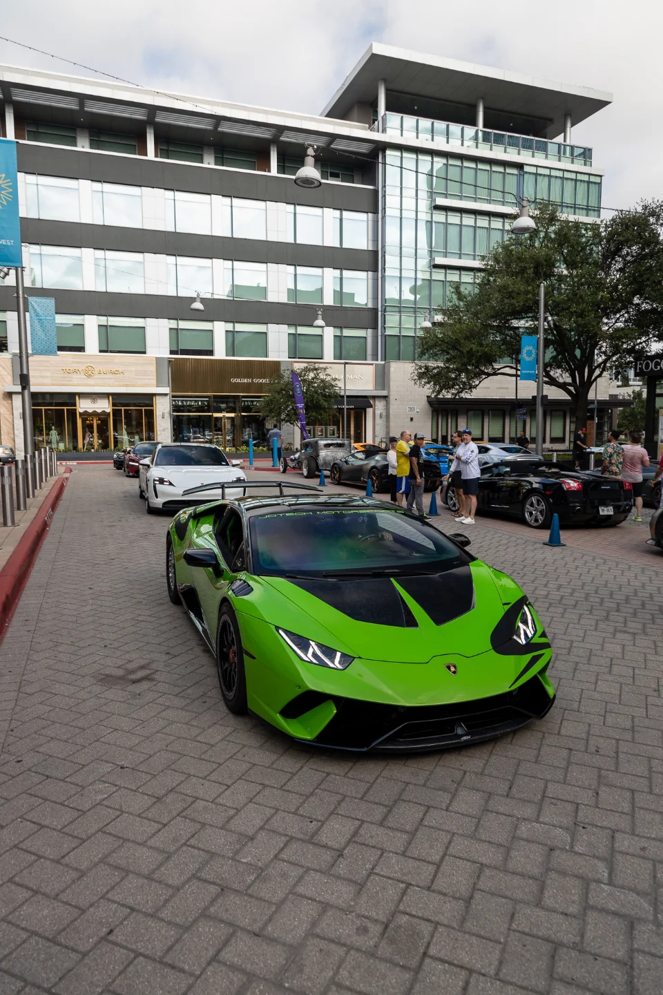 A vibrant green Lamborghini Huracan with black accents parked on a brick street at a car event, with people and other luxury cars in the background.