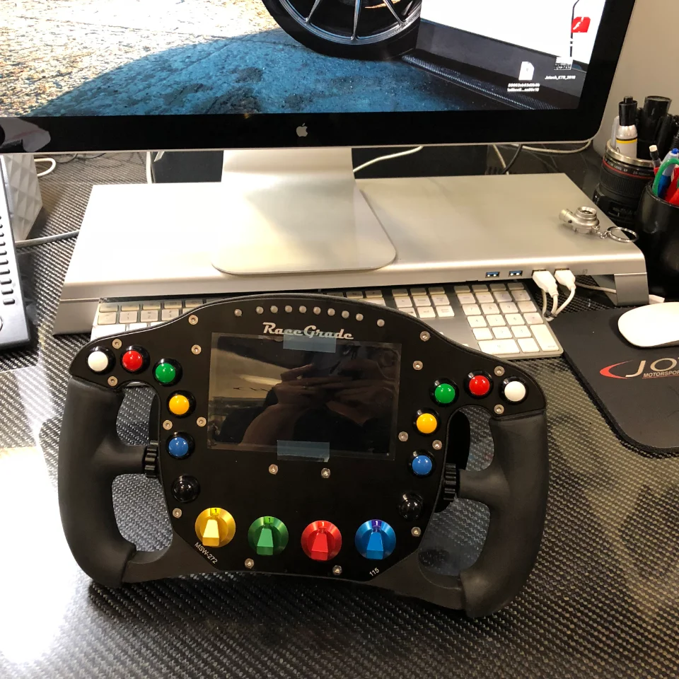 RaceGrade racing steering wheel with multiple buttons and a screen on a carbon fiber desk in front of an iMac.