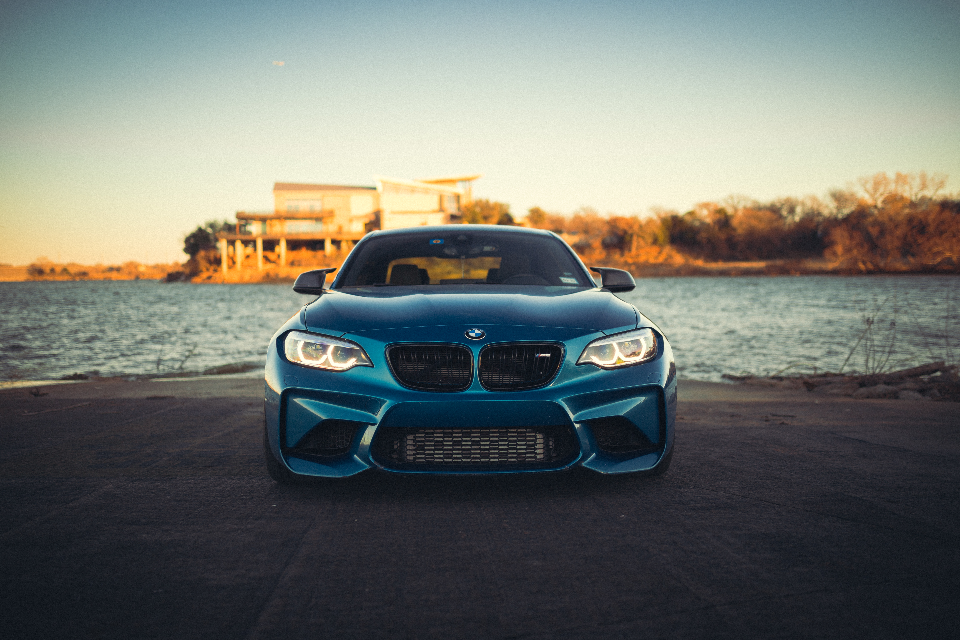 A blue BMW M2 performance car parked by a lake at sunset, viewed from the front with its headlights on.