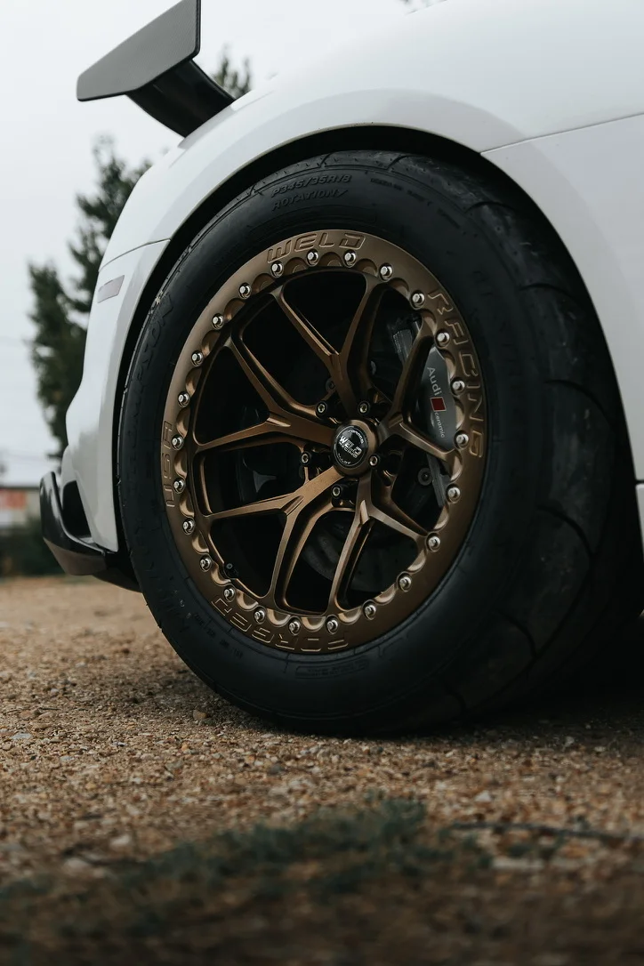 Close-up of a white Audi's rear wheel with bronze Weld Racing wheels, drag radial tires, and a carbon fiber wing.