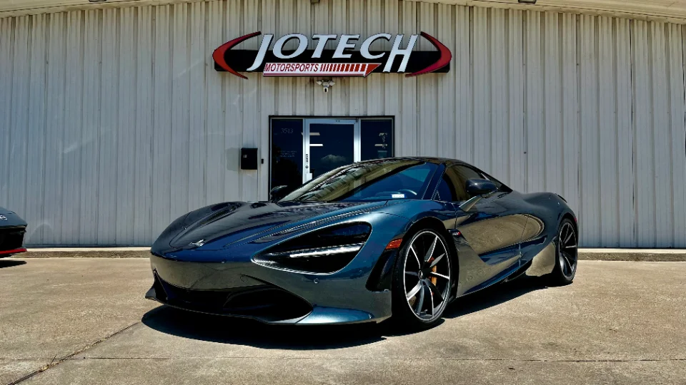 A dark grey McLaren 720S supercar parked in front of the Jotech Motorsports shop building on a sunny day.