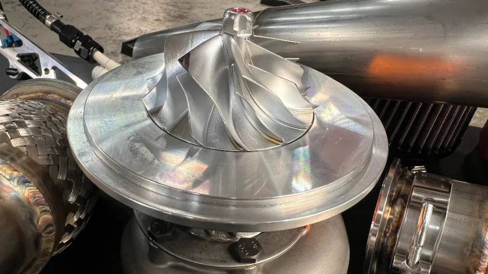 A close-up shot of various performance car parts including a polished turbocharger compressor wheel, an exhaust flex pipe, and an air filter.