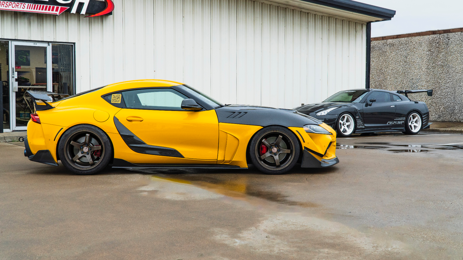A yellow modified Toyota Supra with a carbon fiber hood and a black Nissan GT-R with a large rear wing parked outside Jotech Motorsports.