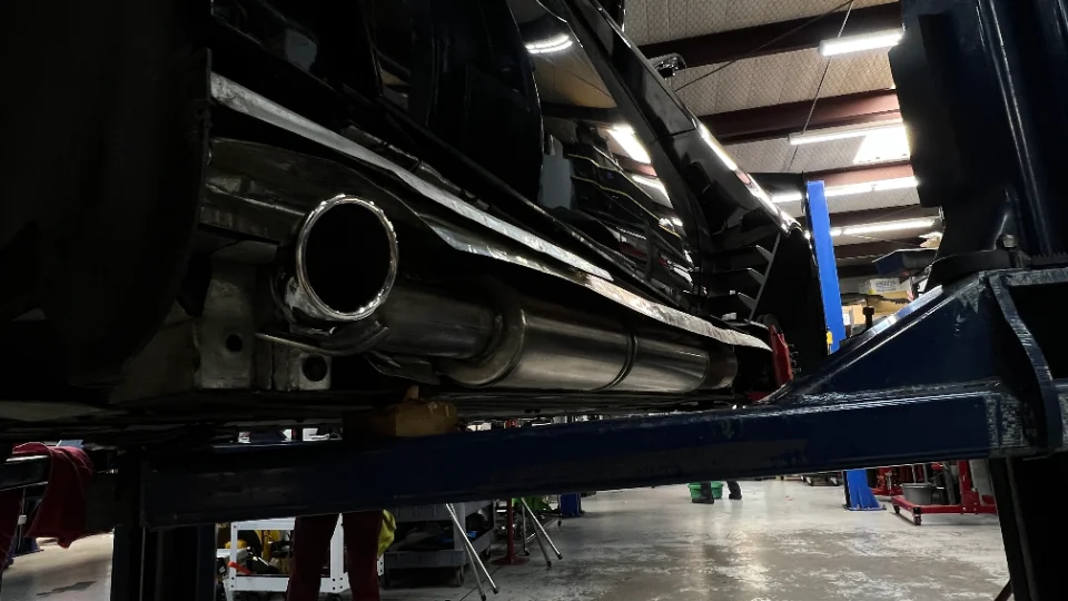 Underside view of a black car on a lift, showcasing a newly installed aftermarket exhaust system in a performance shop.