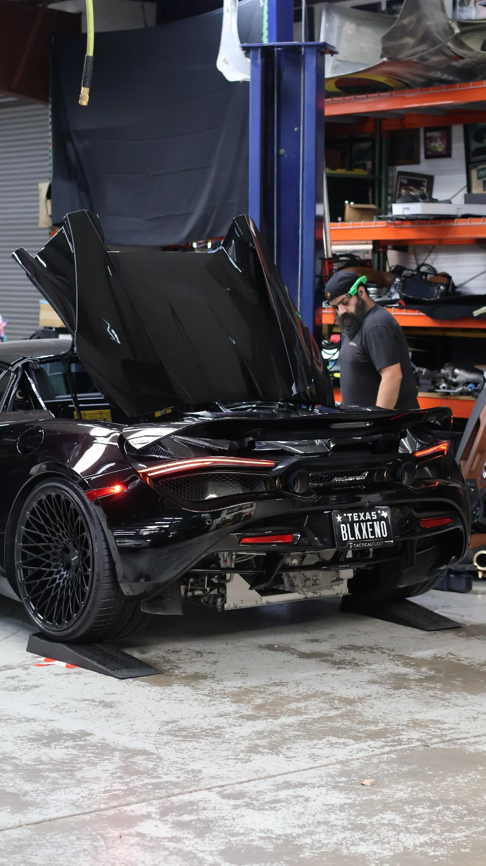 A black McLaren 720S with its engine bay open, being worked on by a mechanic in a performance shop.