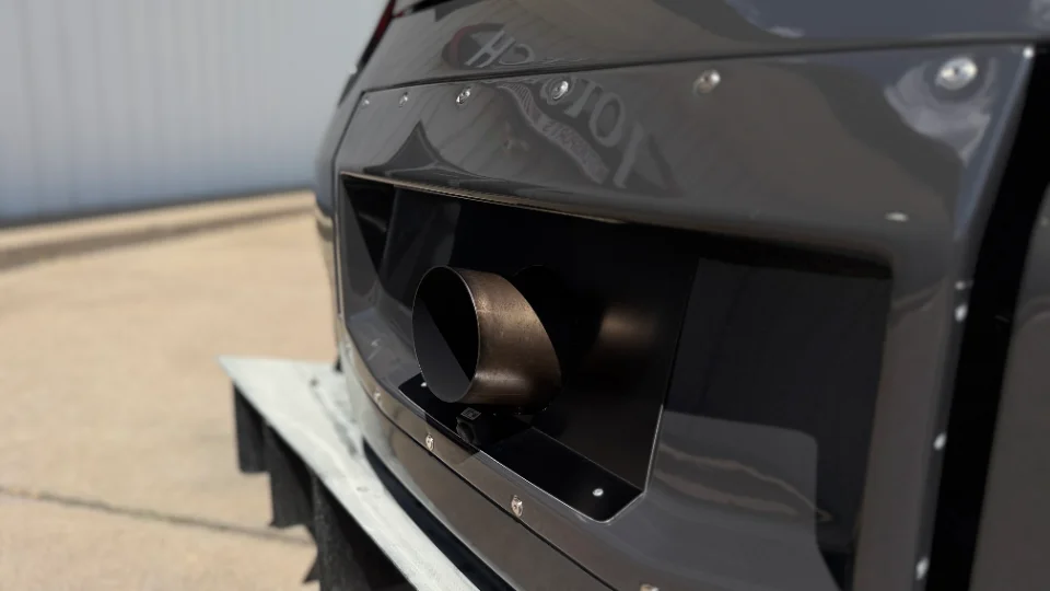 Close-up of a dark grey car's rear bumper featuring a large, custom exhaust tip and widebody fender with visible rivets.