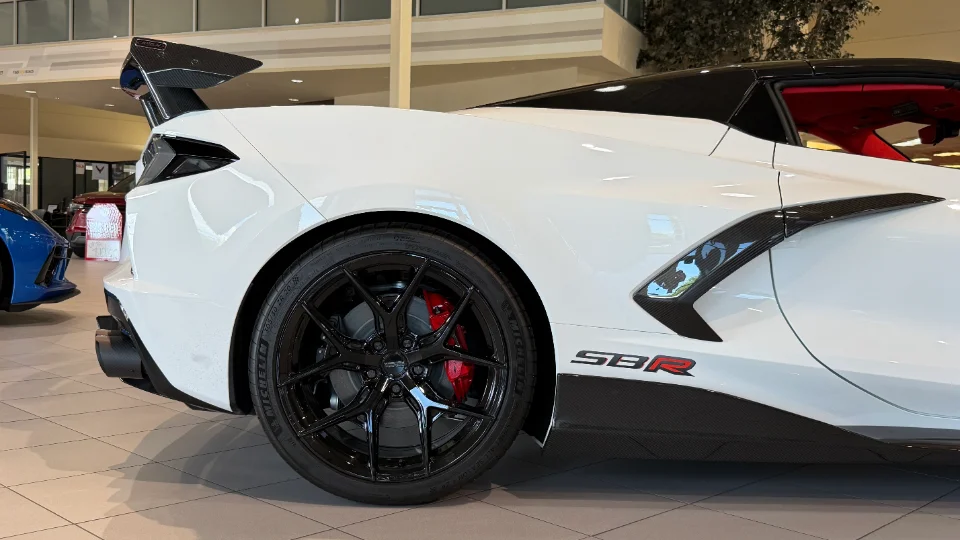 A white Chevrolet Corvette C8 with black aftermarket wheels, red brake calipers, and a large carbon fiber rear wing in a showroom.