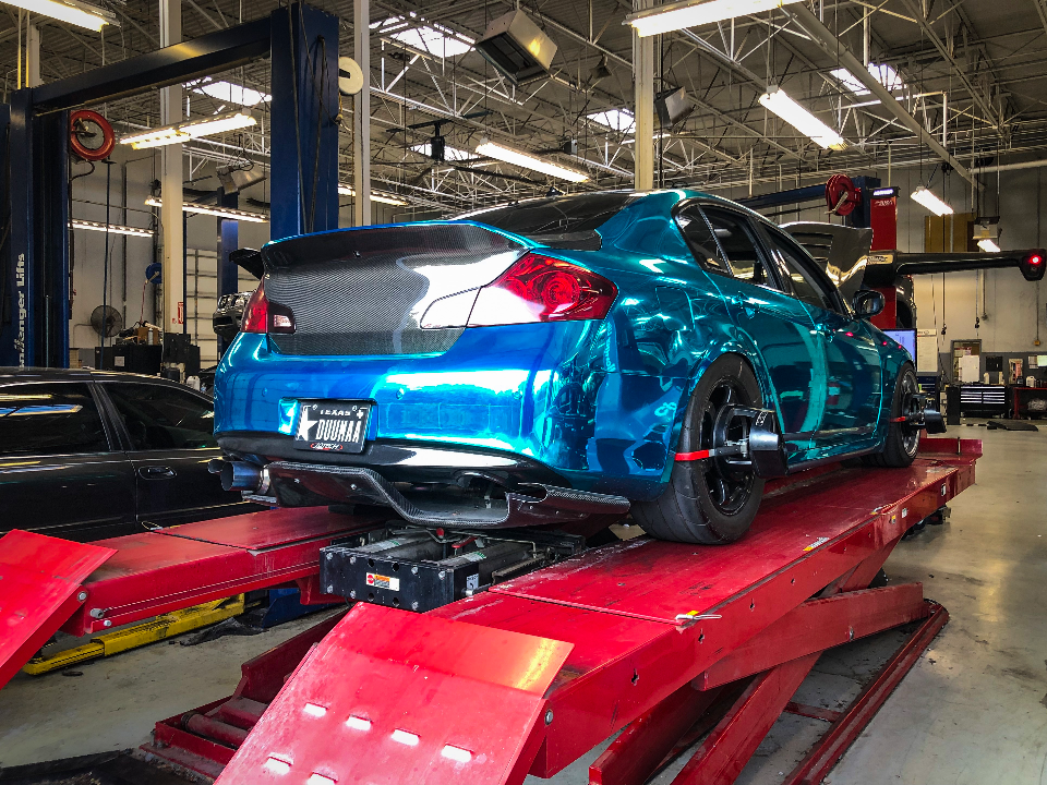 A blue chrome-wrapped Infiniti G37 coupe on an alignment rack in a performance shop.
