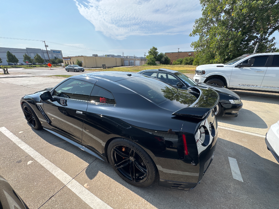 A black Nissan GT-R with aftermarket wheels parked outdoors on a sunny day, showing its sleek rear and side profile.