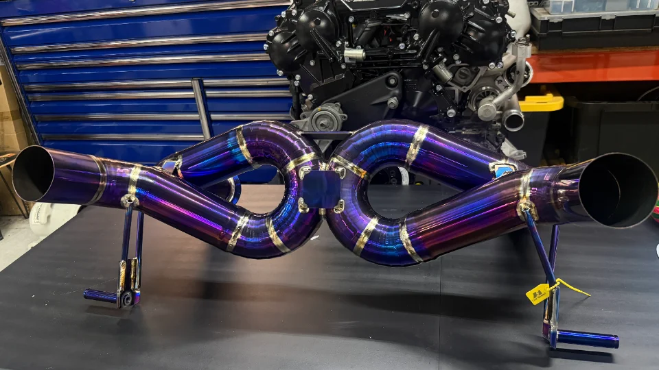 Titanium exhaust system with a vibrant burnt finish, displayed in a performance shop with an engine block in the background.