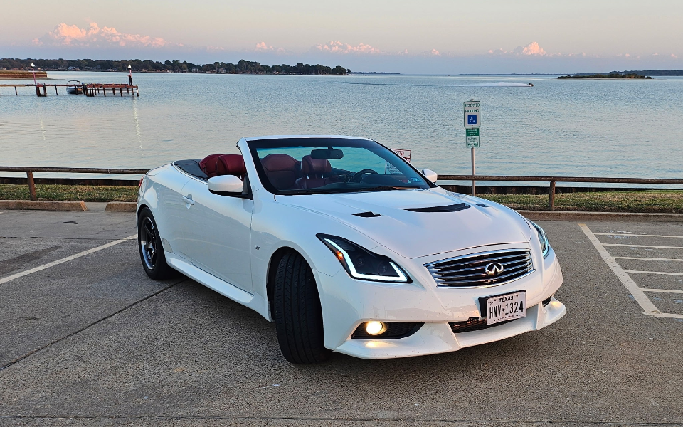A white Infiniti G37 convertible with red interior parked by a lake at sunset, showcasing its sleek design.