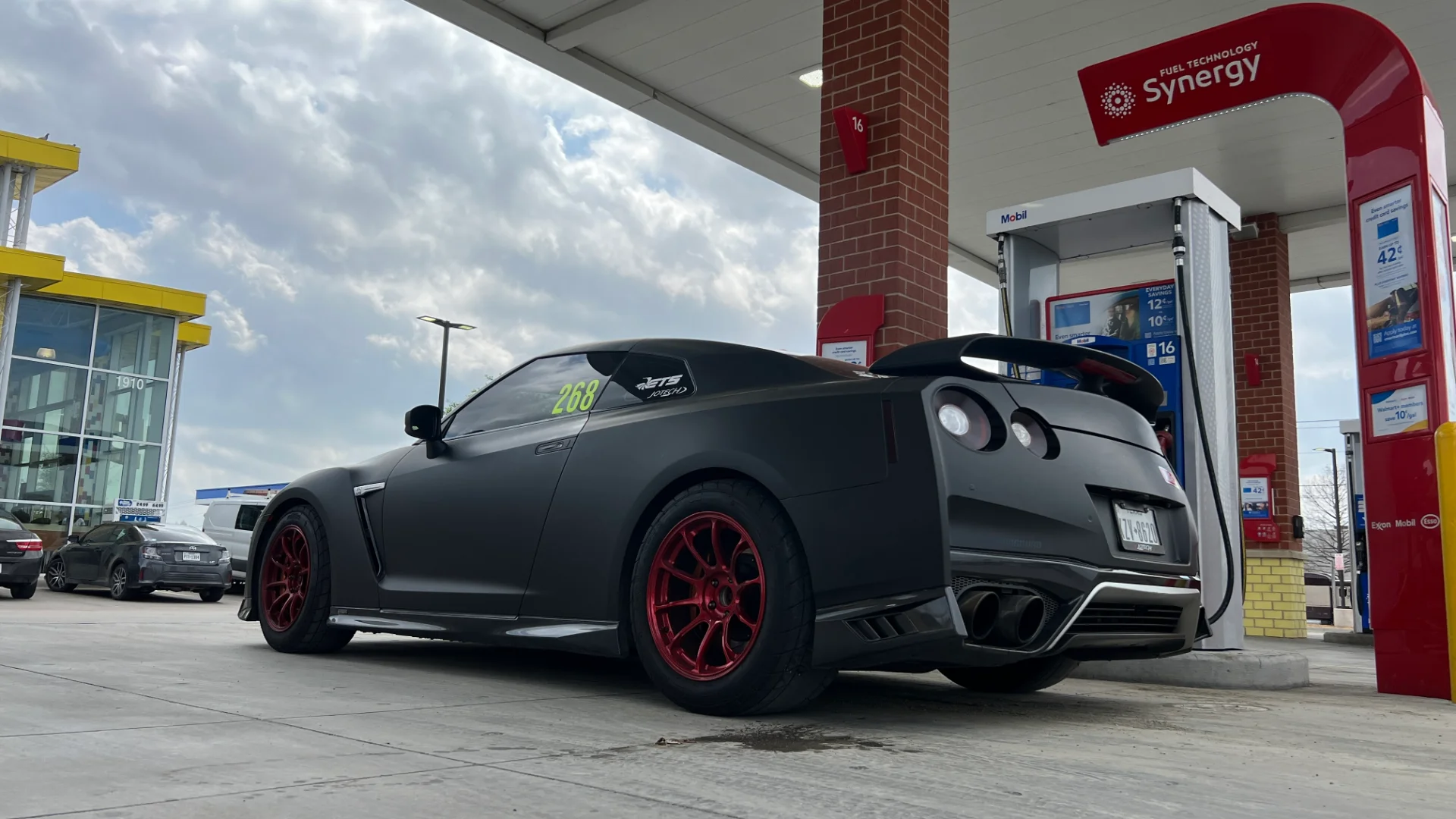 Matte black Nissan GT-R with red aftermarket wheels and a large rear wing parked at a gas station.
