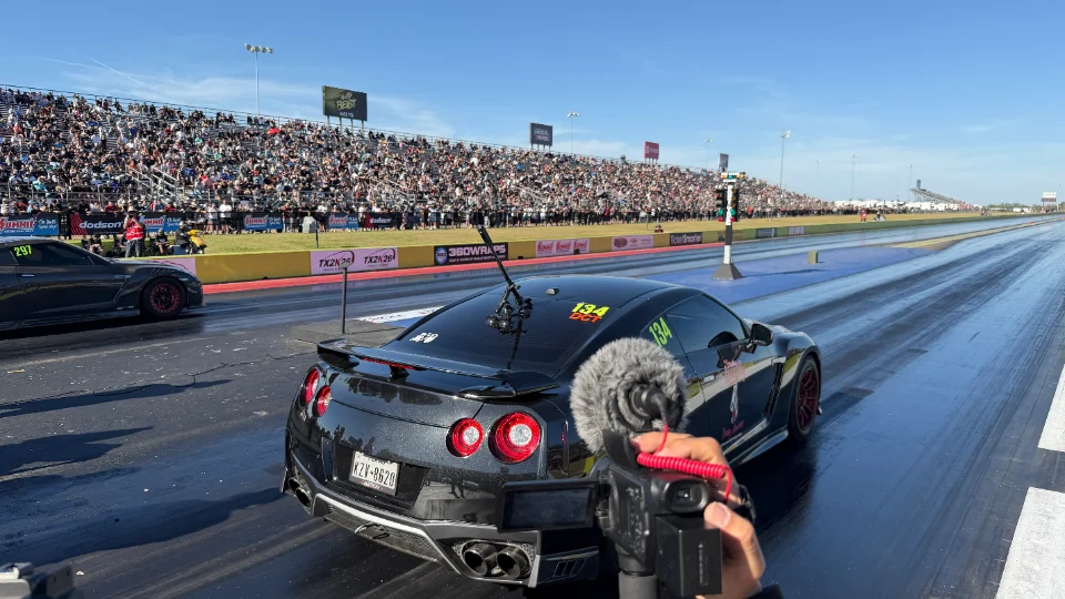 Two Nissan GT-Rs at the starting line of a drag strip with a large crowd in the stands, filmed by a handheld camera.