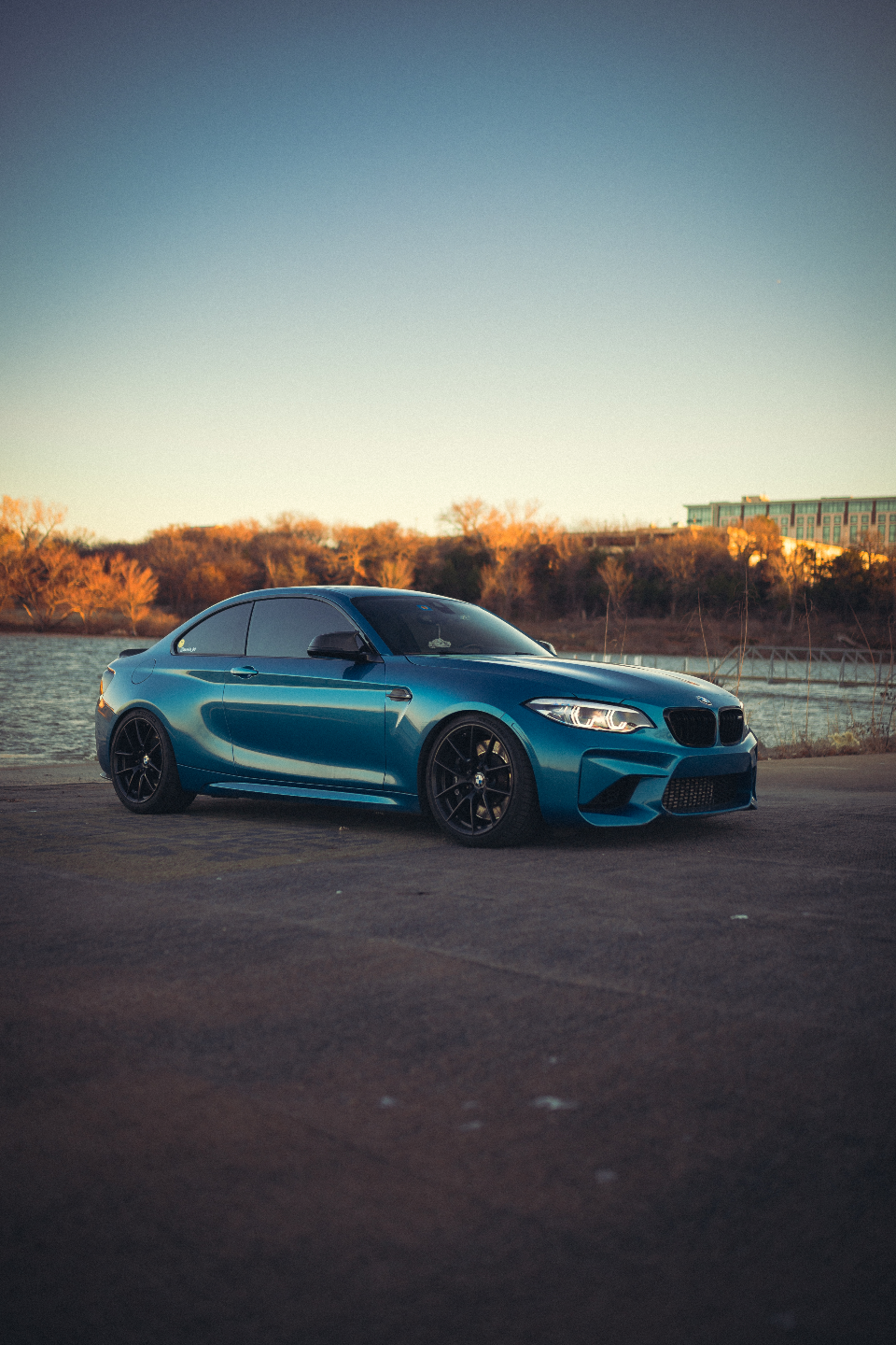 Blue BMW M2 coupe with black wheels parked by a lake at sunset.
