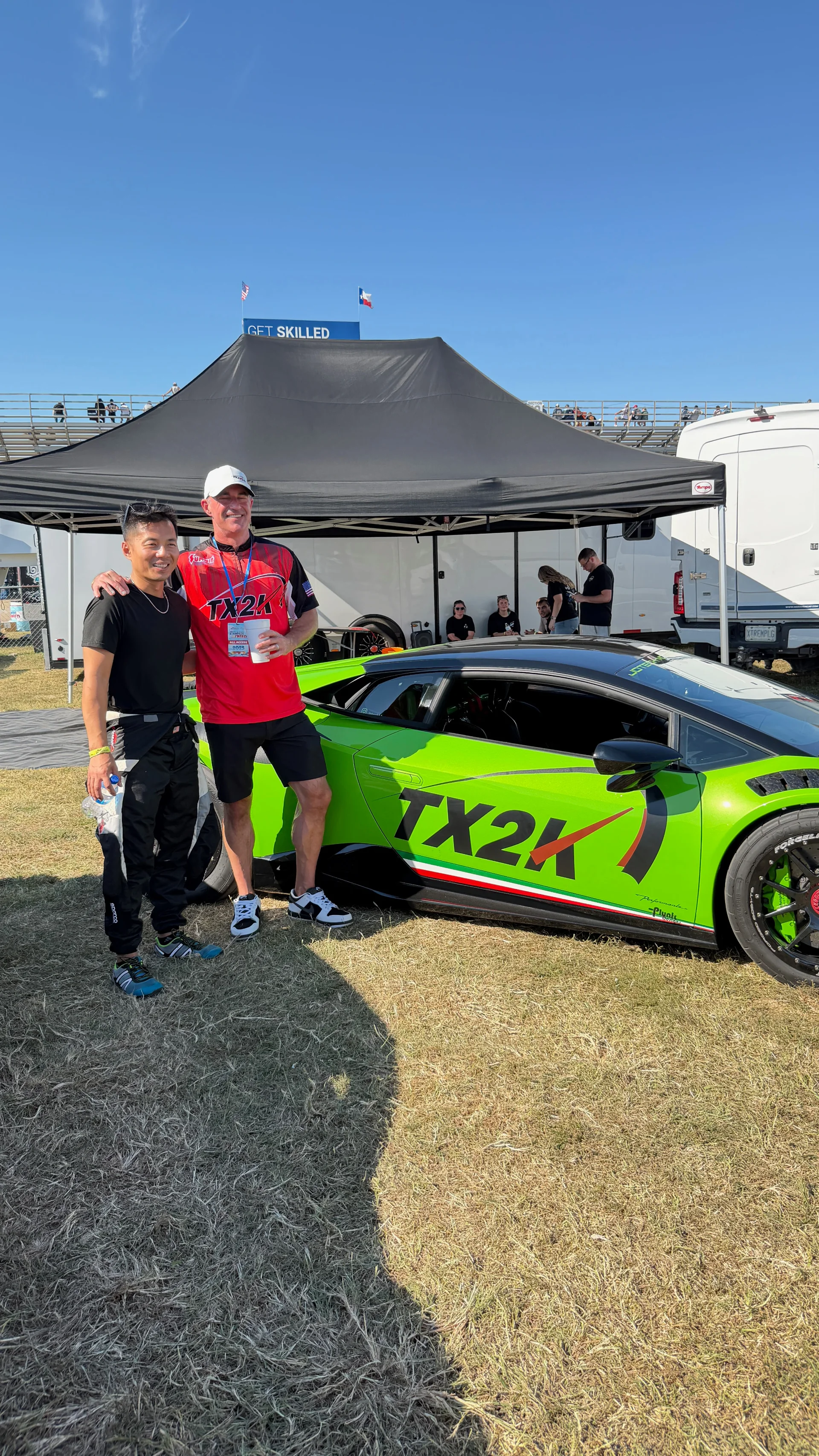 A vibrant green Lamborghini Huracan with TX2K livery is parked on grass at an outdoor event, with two men standing beside it.