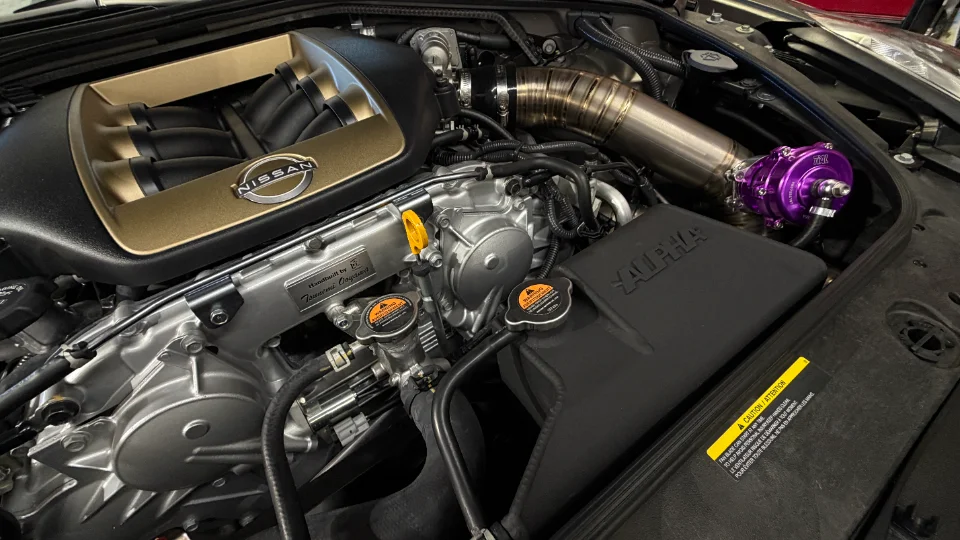 Close-up of a modified Nissan GT-R VR38DETT engine bay featuring an aftermarket intake manifold, boost piping, and a purple blow-off valve.
