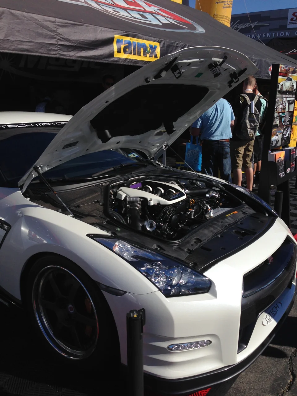 White Nissan GT-R with its hood open, showcasing a modified engine bay, displayed under a RainX tent at an event.