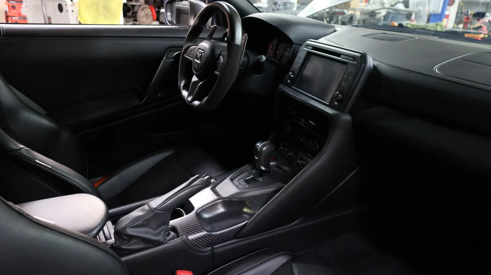 Interior view of a Nissan GT-R featuring the steering wheel, dashboard, center console with carbon fiber trim, and black leather seats.