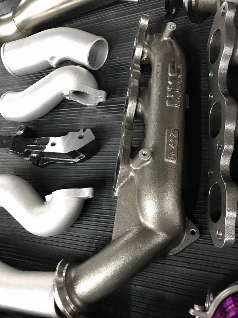 Close-up of an HKS turbo manifold and various silver boost pipes laid out on a dark surface.