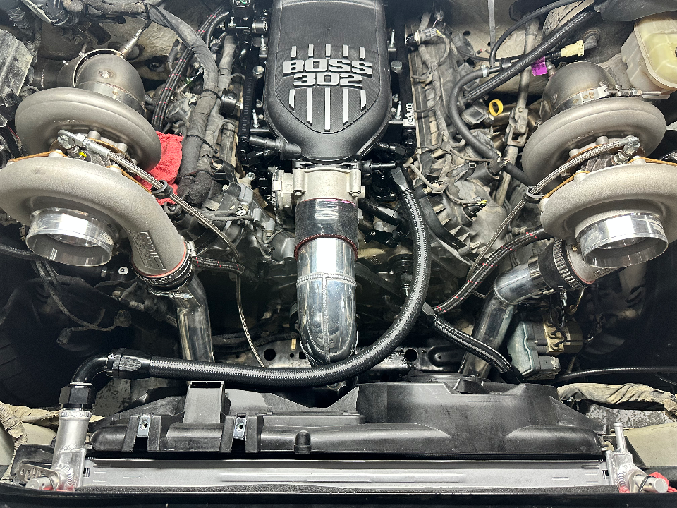 Close-up view of a custom twin-turbo engine bay featuring a Boss 302 intake manifold and intricate boost piping.