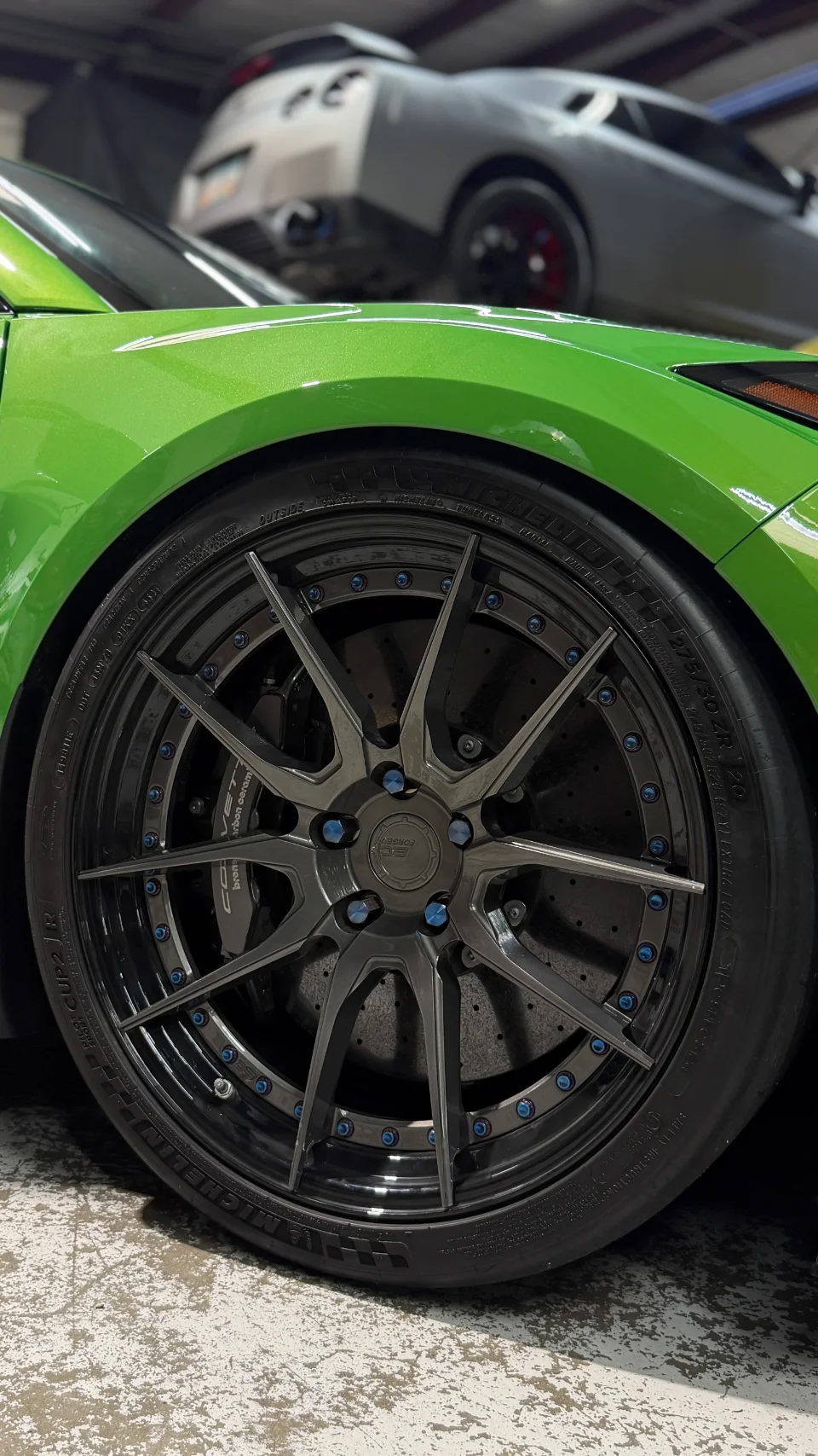 Close-up of a bright green car's front wheel, featuring dark forged wheels with blue lug nuts, Michelin tires, and Brembo brakes in a shop.