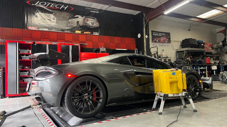 A grey McLaren 570S with a carbon fiber wing strapped to a dynamometer at Jotech Motorsports for performance tuning.