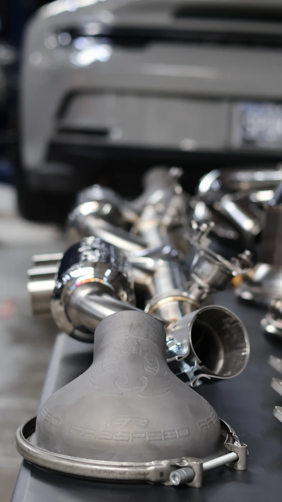 Close-up of Fabspeed GT3 exhaust system components, including polished mufflers and piping, with a blurry sports car in the background.