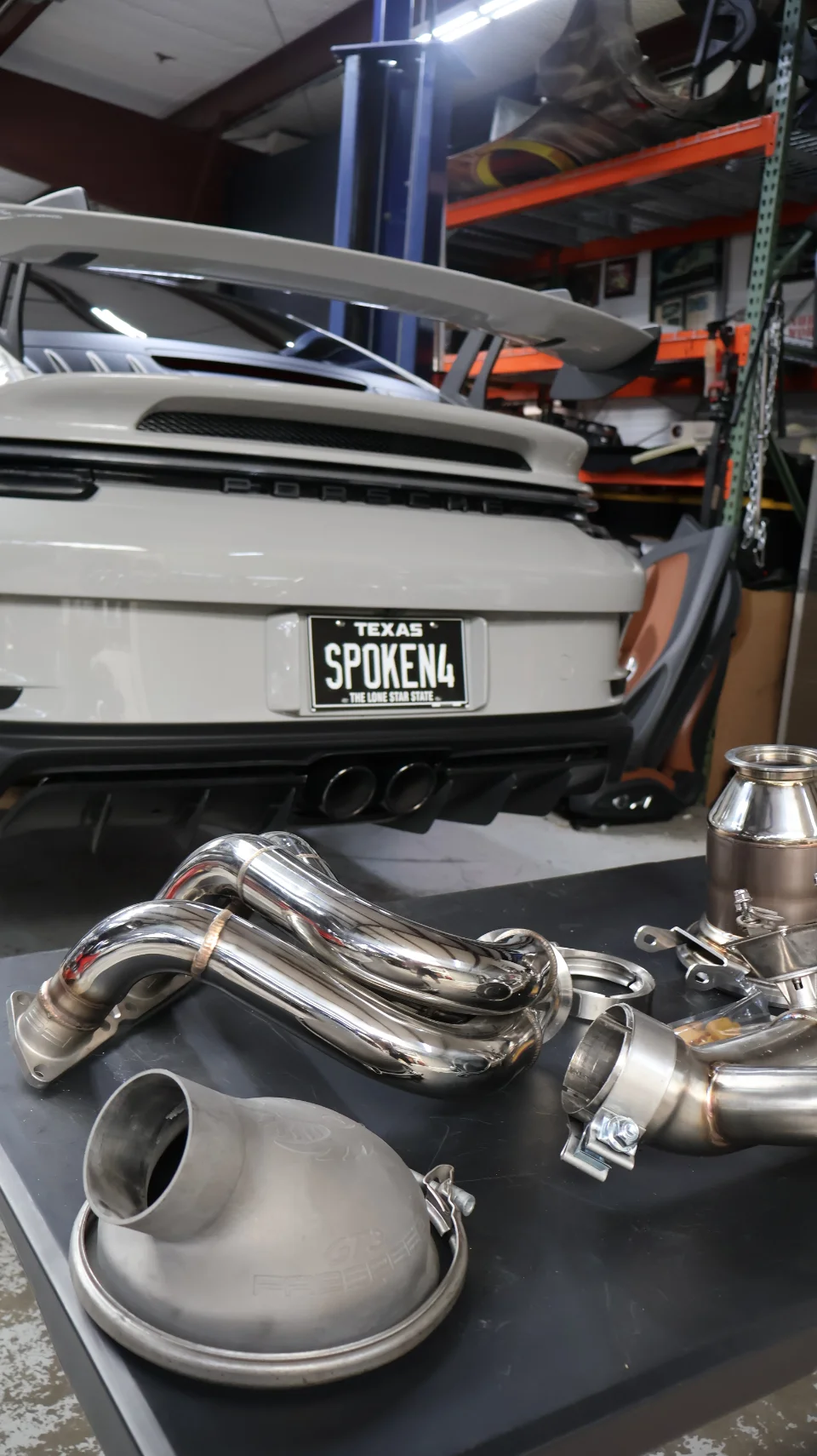 A white Porsche 911 GT3 on a lift in a shop with aftermarket exhaust headers and components on a table in the foreground.